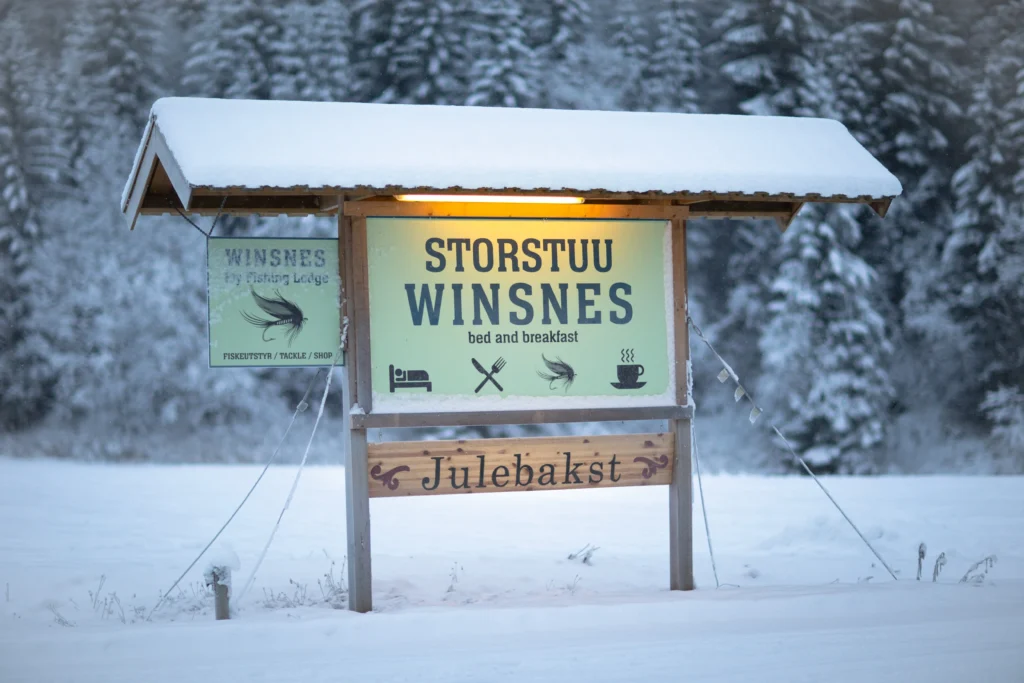 A snow-blanketed sign for "Storstuu Winsnes Bed and Breakfast," adorned with dining and lodging icons, stands elegantly against a wintry forest backdrop, offering a perfect retreat following the enchanting Julemesse festivities.