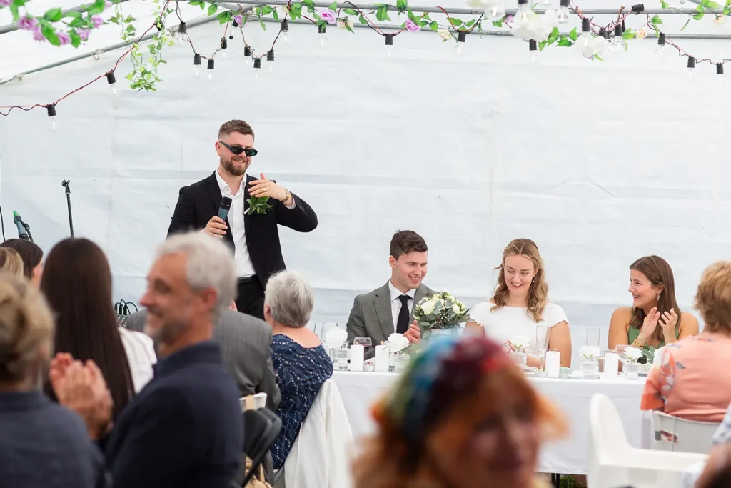 At a charming wedding reception in Selskapslokaler Gauldal, a man in sunglasses speaks into the microphone. Guests seated at the long table, including the smiling bride and groom, listen intently to his speech.