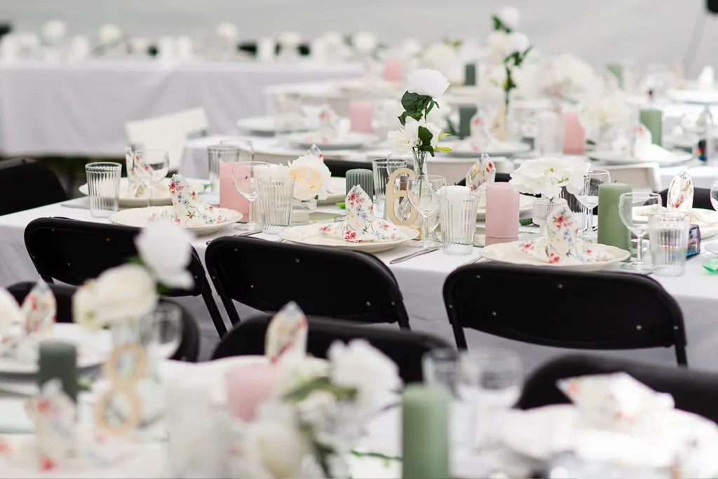 Tables are elegantly set for an event in the charming selskapslokaler Gauldal, featuring white tablecloths, black chairs, floral plates, pastel candles, and stunning white flower centerpieces.