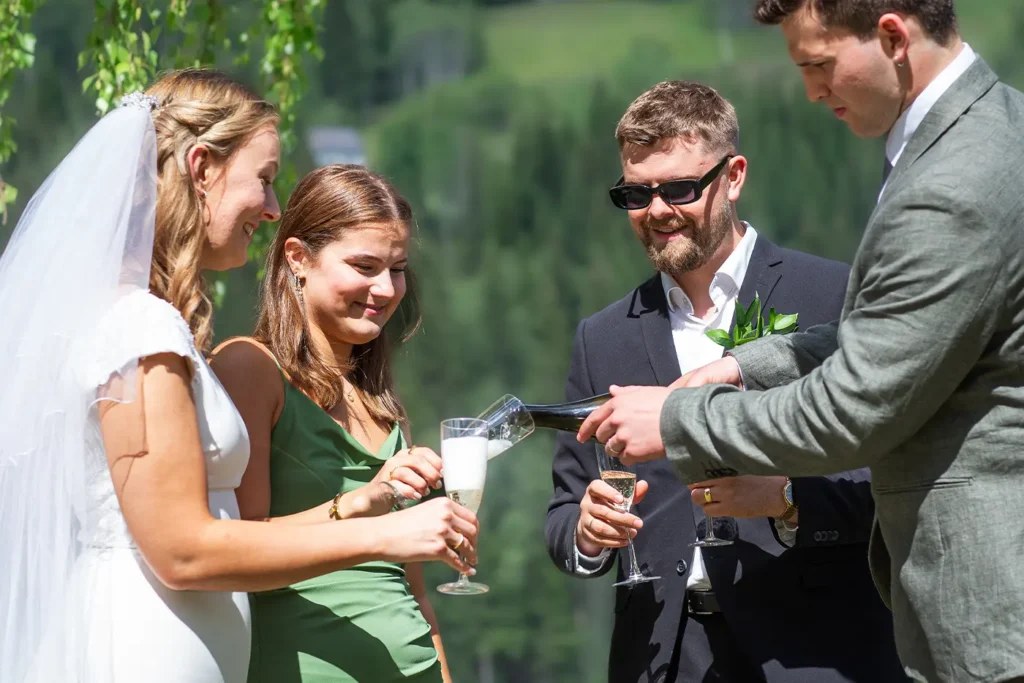 A bride and three guests toast outdoors at Storstuu Winsnes. One person pours champagne into a glass, with lush greenery creating a picturesque background.